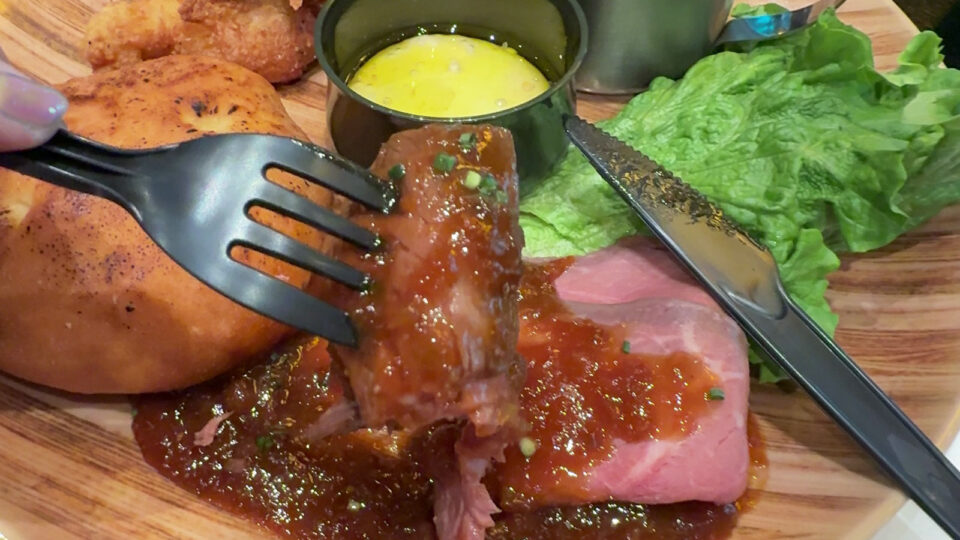 ユニバーサル・スタジオ・ジャパンのモンスターハンターワイルズ・コラボプレート。フォークで持ち上げた厚切りローストビーフのシズル感あふれるアップ。