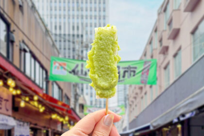 仙台朝市を背景に、串に刺さった鮮やかなずんだ餅を持つ手元。仙台日帰り観光の始まりにぴったりな食べ歩きグルメ。