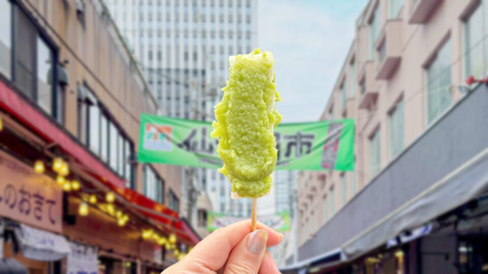 仙台朝市を背景に、串に刺さった鮮やかなずんだ餅を持つ手元。仙台日帰り観光の始まりにぴったりな食べ歩きグルメ。